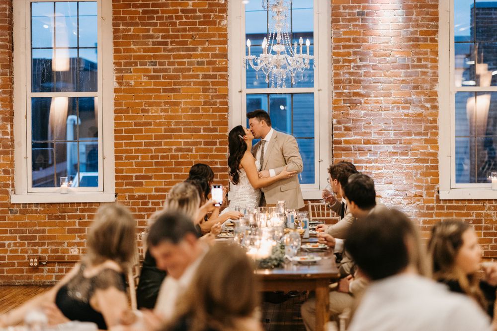 Bride and groom share a kiss at candlelit reception table in rustic brick venue.