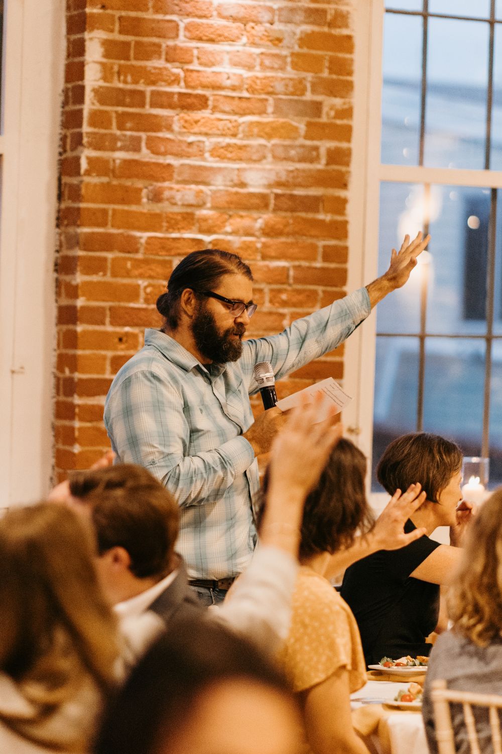 Bearded man in plaid shirt gives heartfelt wedding toast, gesturing with raised hand.
