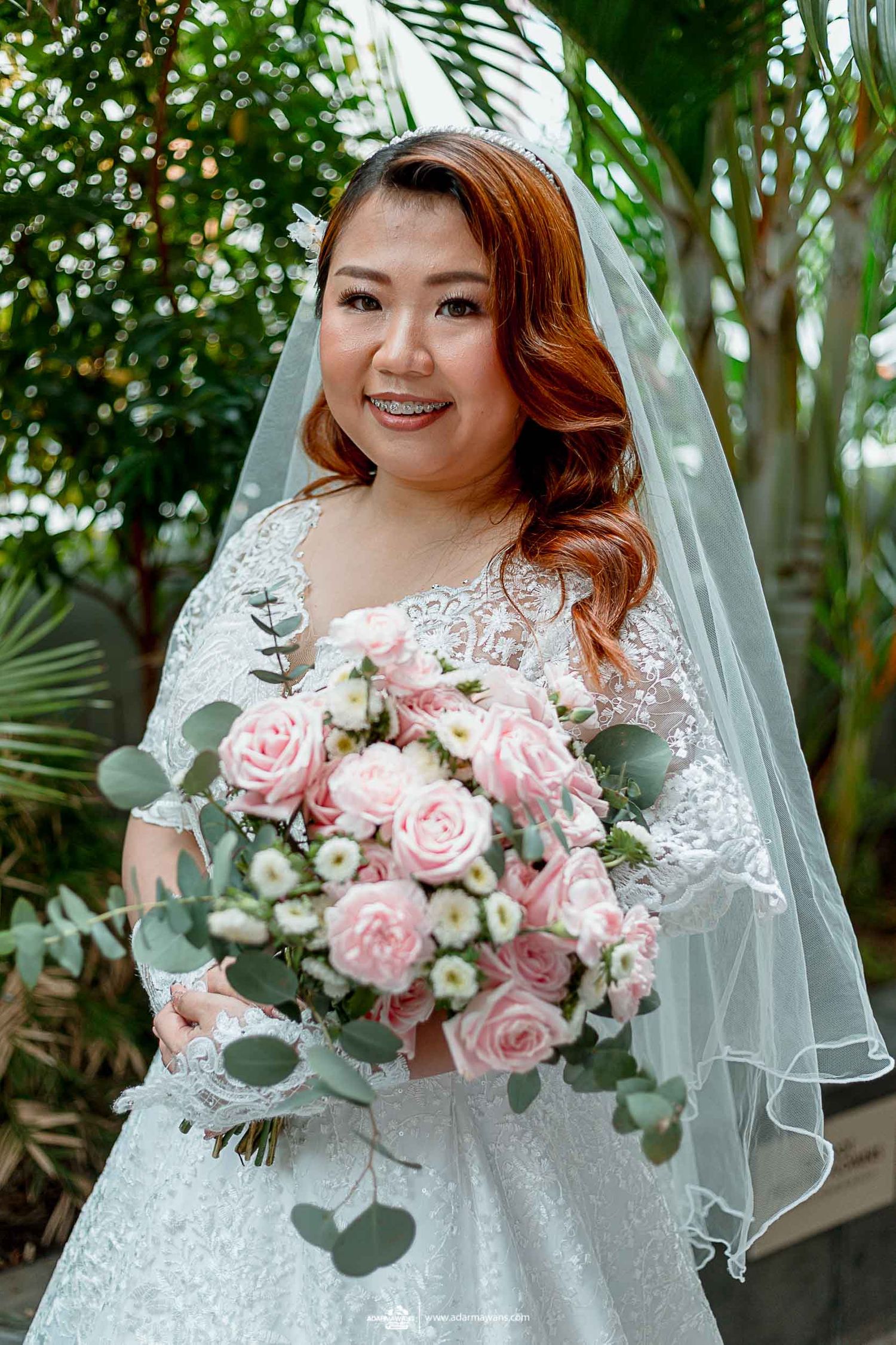 Bride in lace wedding dress holds beautiful pink rose and eucalyptus bouquet in garden setting.