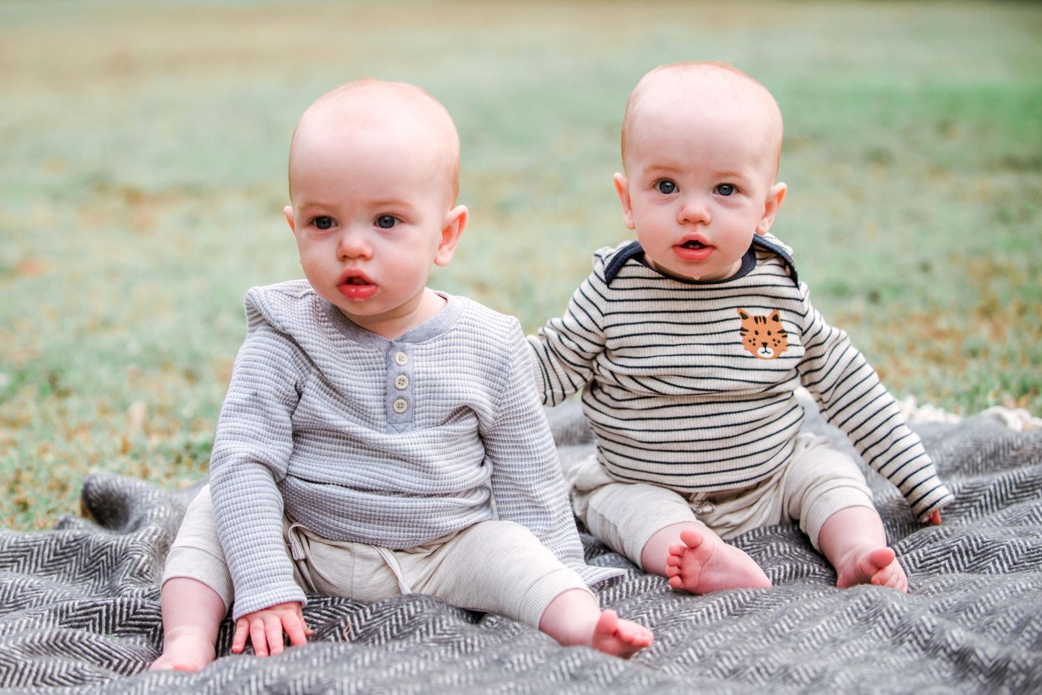 Twin babies in matching grey outfits sitting on blanket during outdoor photo session.