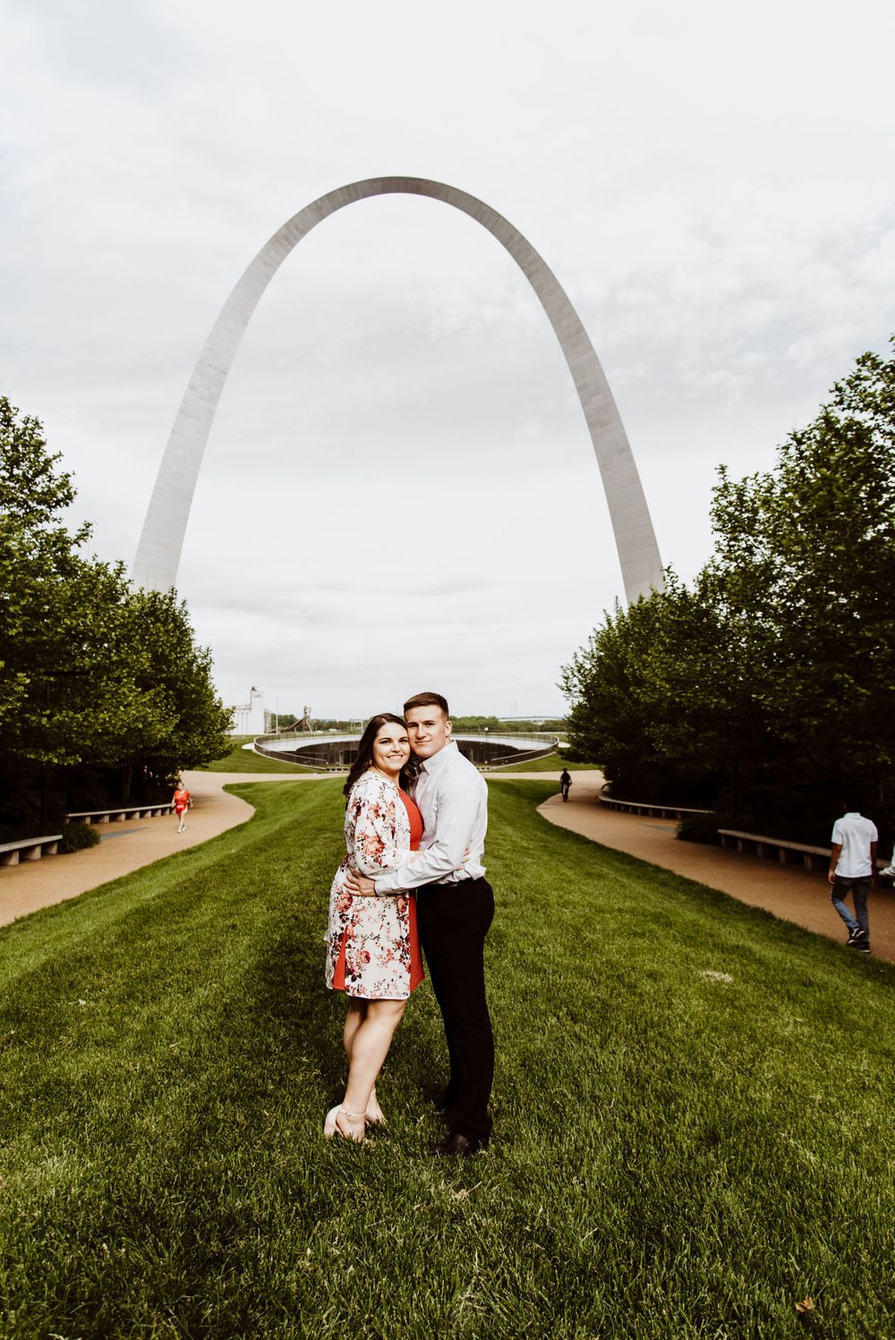 Couple poses beneath the iconic St. Louis Gateway Arch on a grassy hill.