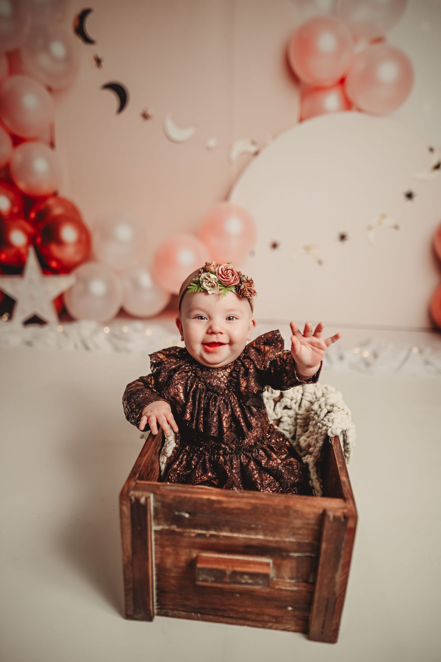 Baby in brown dress sitting in rustic wooden box with pink balloon backdrop.