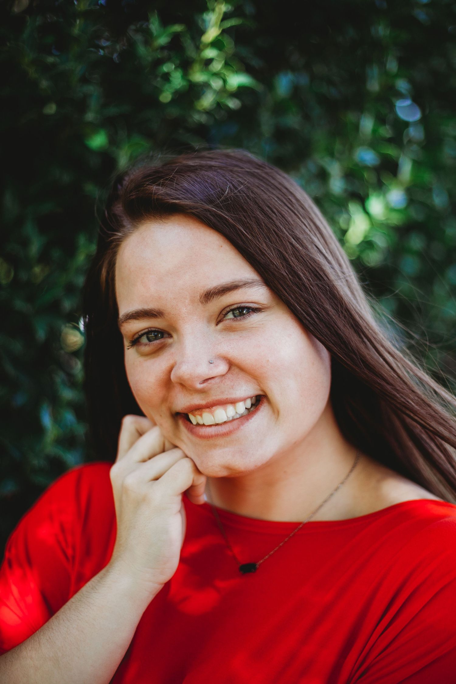 A portrait of someone wearing a red shirt smiling warmly against a green foliage background.