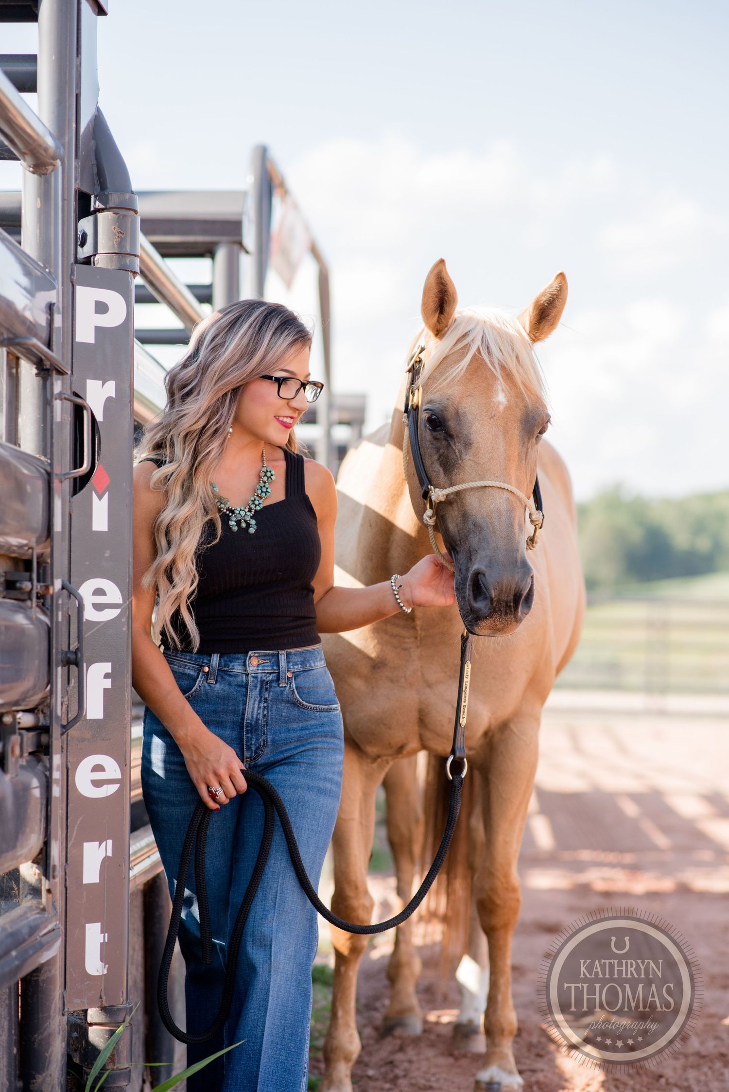 A palomino horse stands beside someone in casual attire near metal horse stalls.