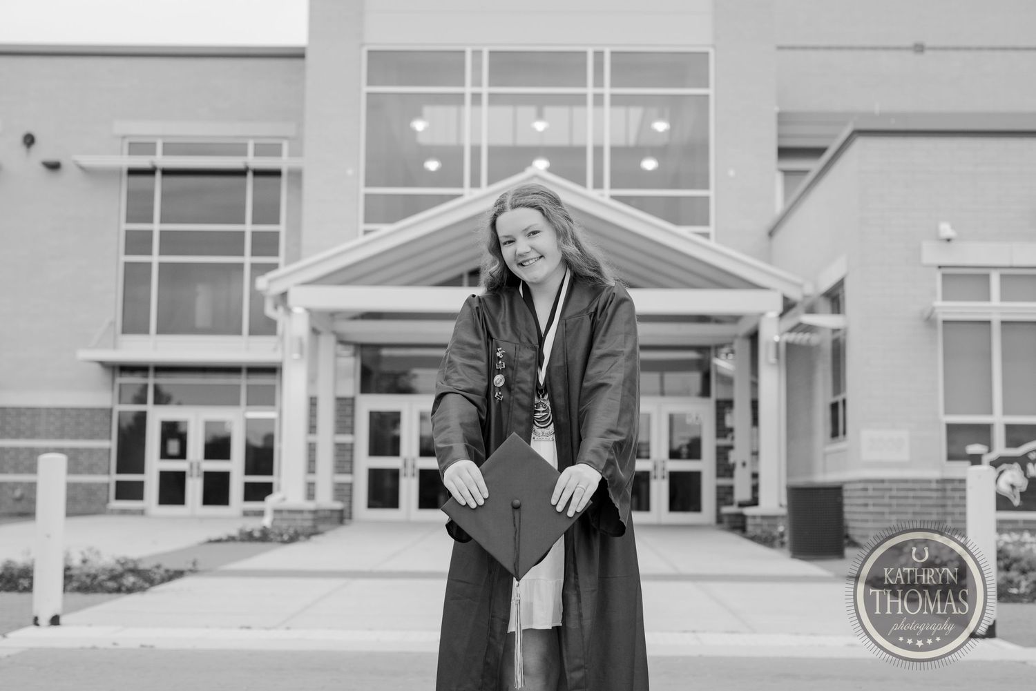 Black and white photo of graduate holding cap in front of academic building.