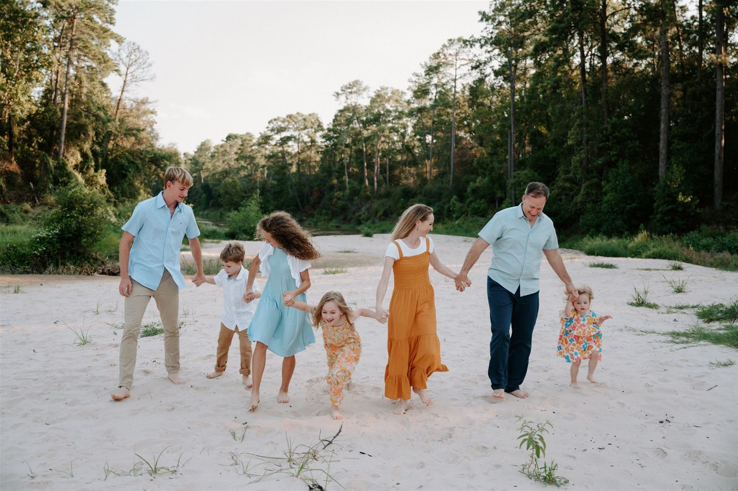 A group in coordinated blue and orange outfits holds hands while walking on a sandy beach path.