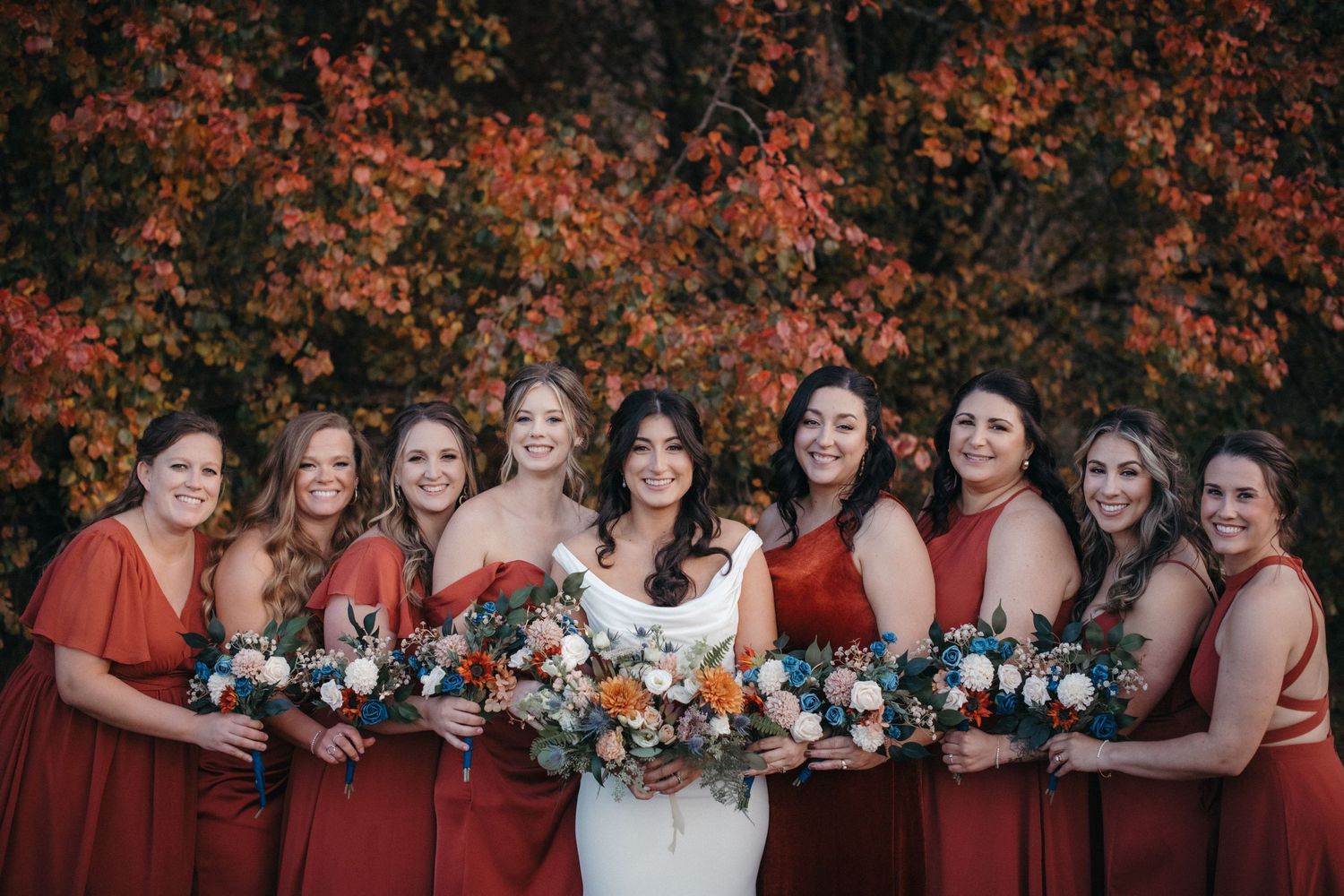 Bridesmaids in rust-colored dresses hold floral bouquets against autumn foliage.