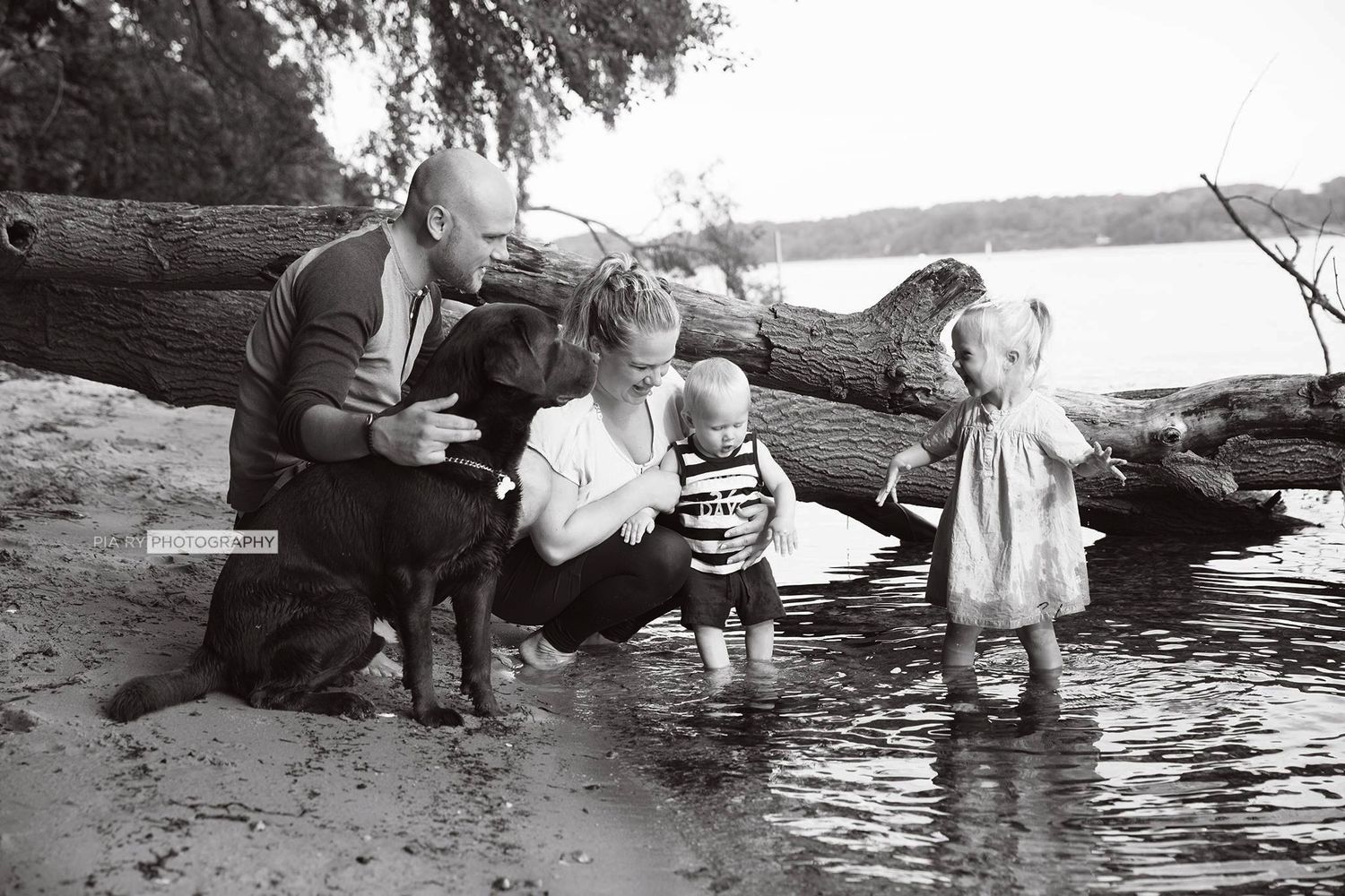 En familie nyder en dag ved vandet, hvor børn leger i det lave vand, mens en voksen sidder på en træstamme.