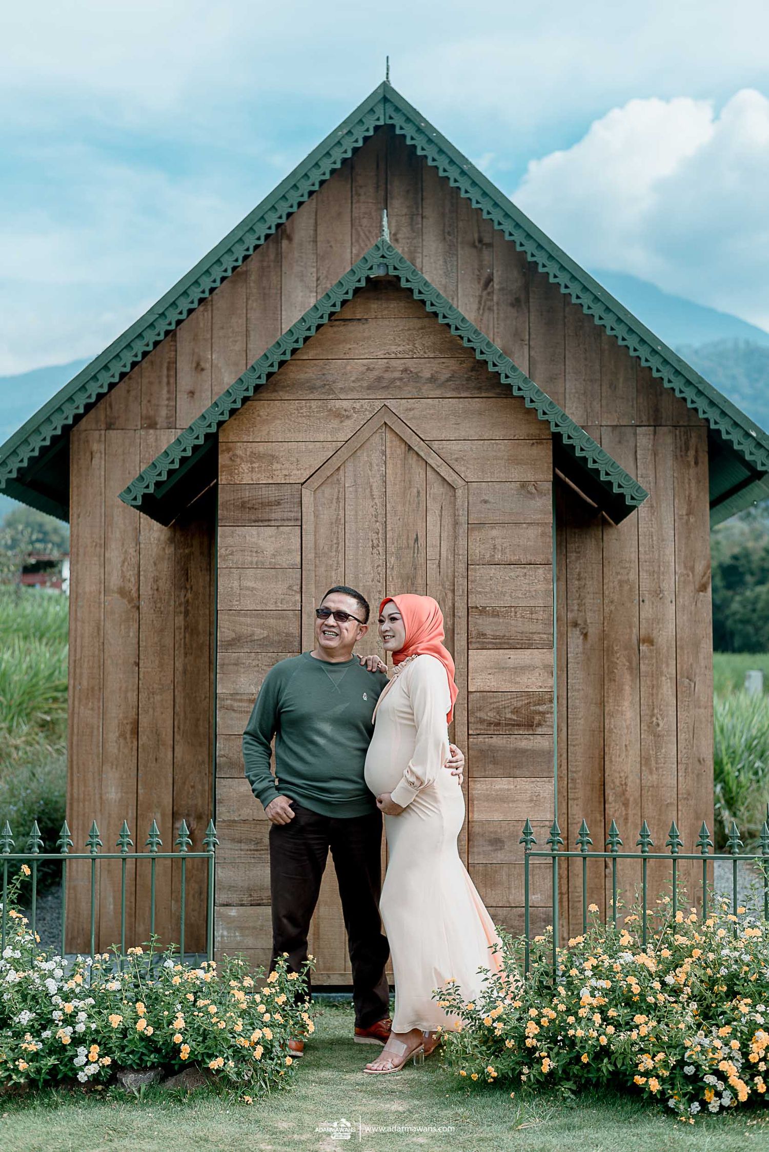Wooden chapel with green trim surrounded by blooming flowers in a mountain setting.