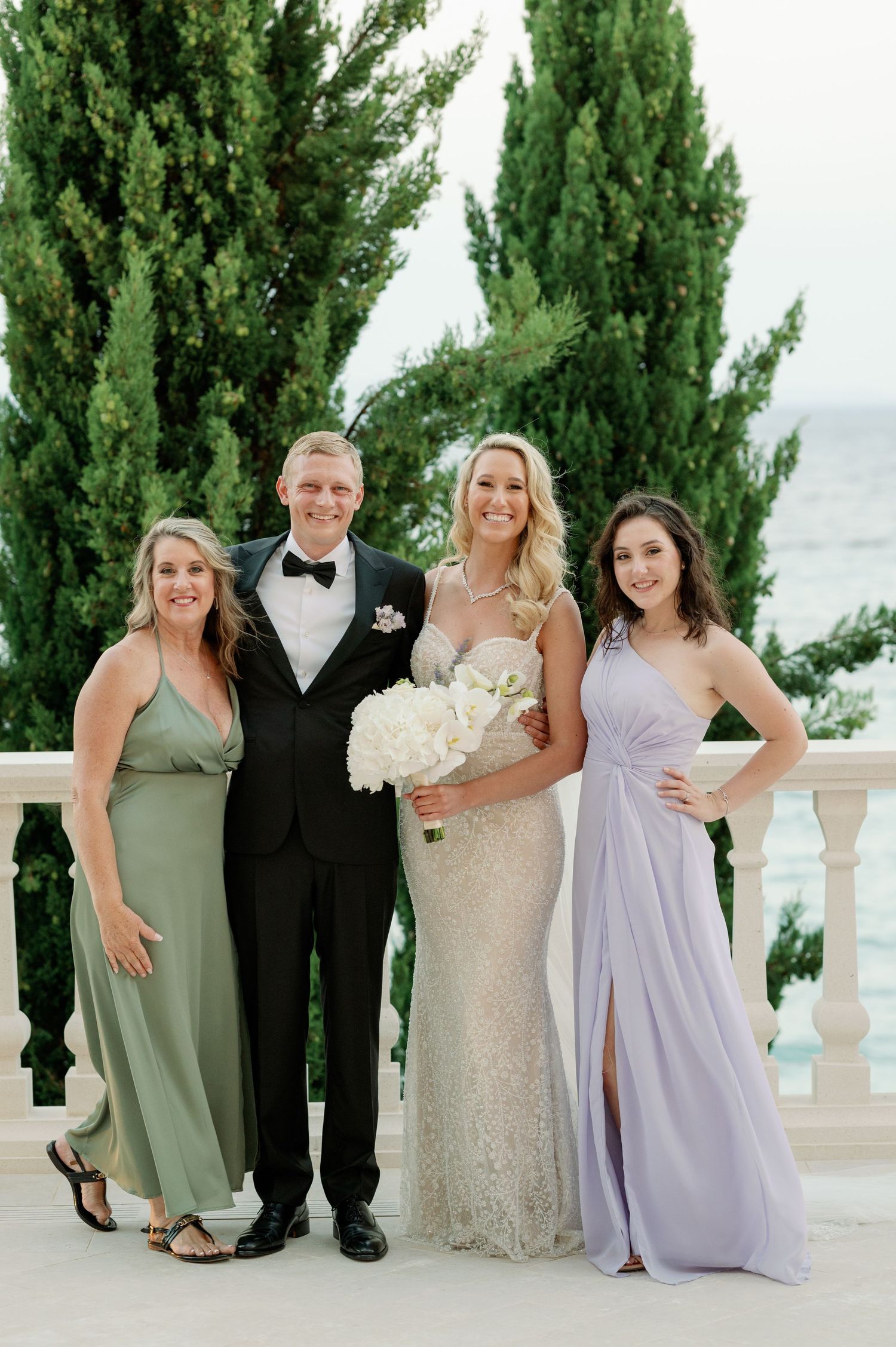 Wedding party poses outdoors with bride in sequined gown and guests in elegant dresses against a scenic backdrop.