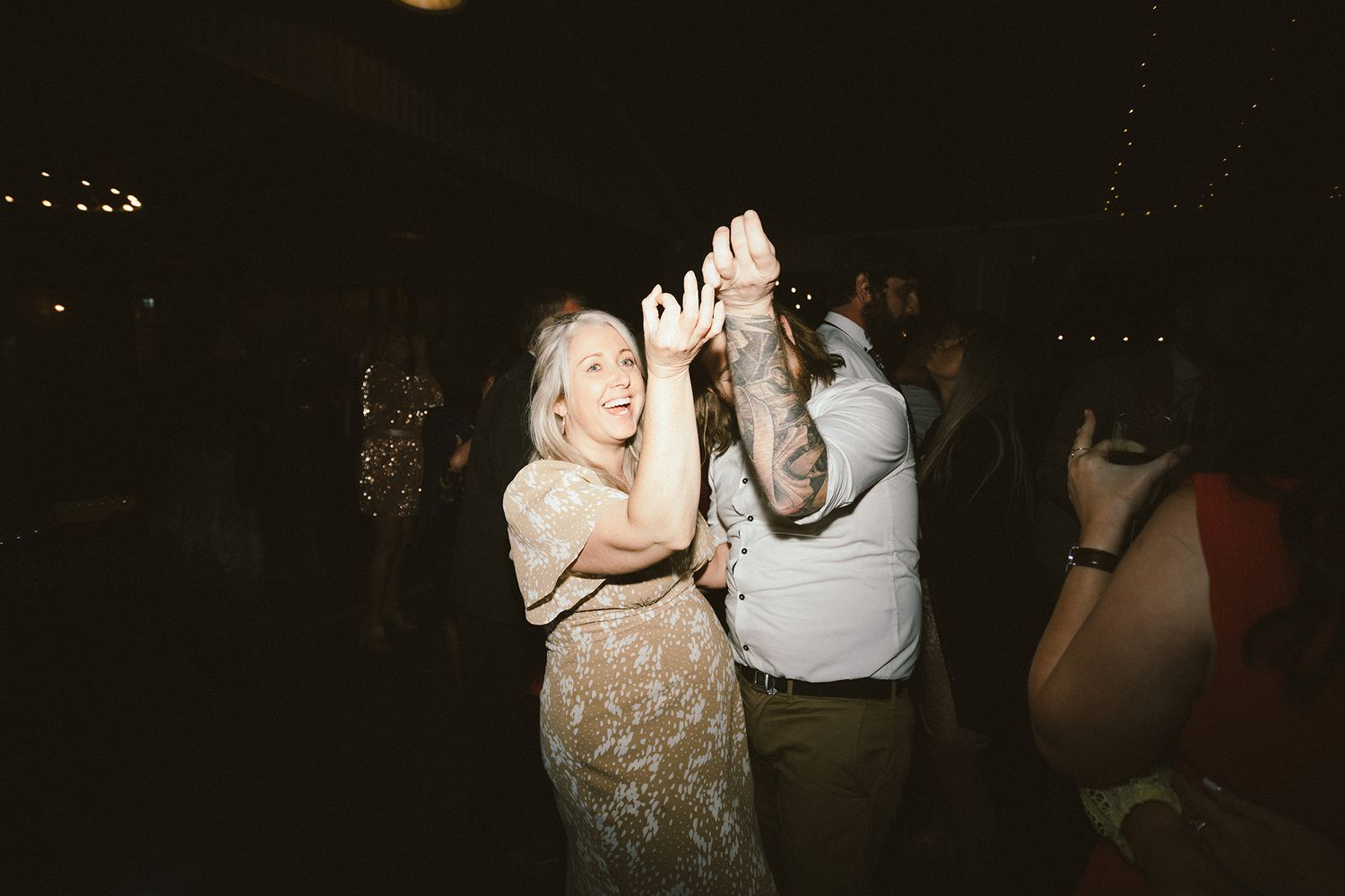 A group of wedding guests dancing together under romantic string lights at an evening celebration.