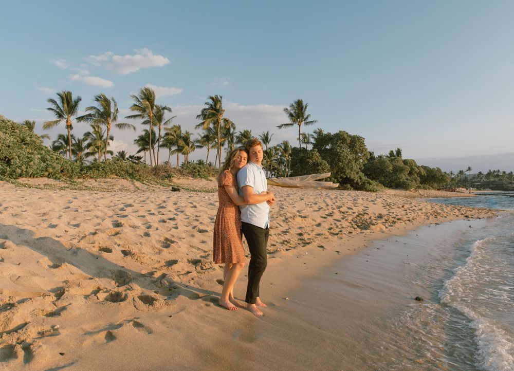 A couple stands together on a sandy Hawaiian beach with palm trees and ocean views.