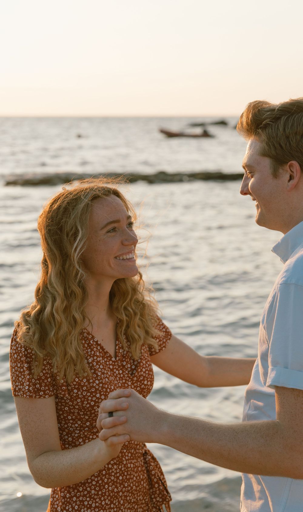 A couple embraces during sunset by the ocean with a boat in the distance.