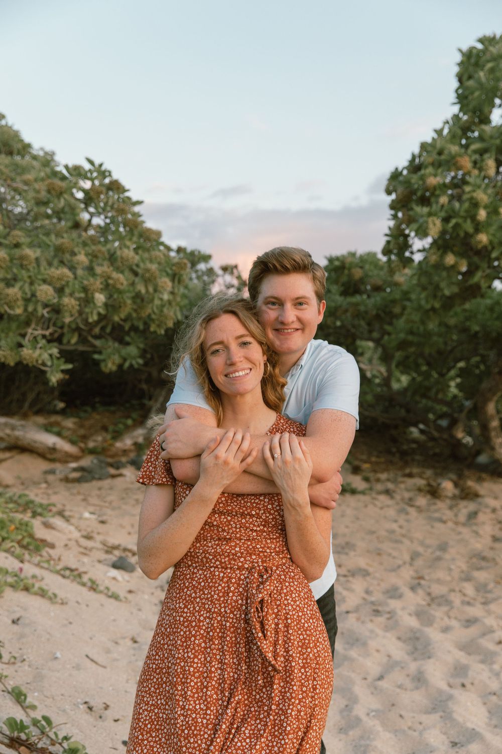 A couple embraces on a beach surrounded by tropical vegetation at sunset.