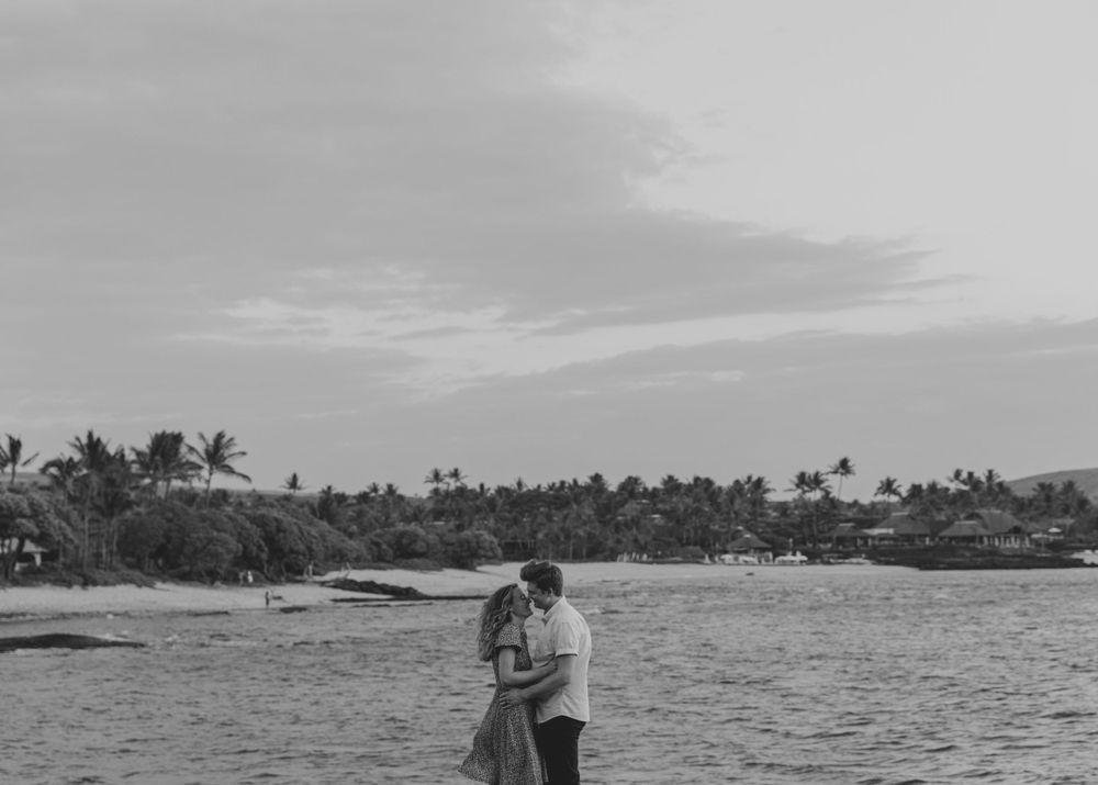 A black and white photo of a couple embracing on a scenic beach with palm trees.