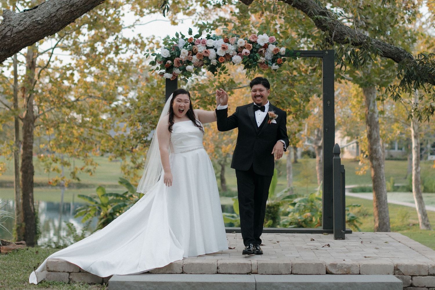 Couple stands under floral arch during autumn outdoor wedding ceremony.