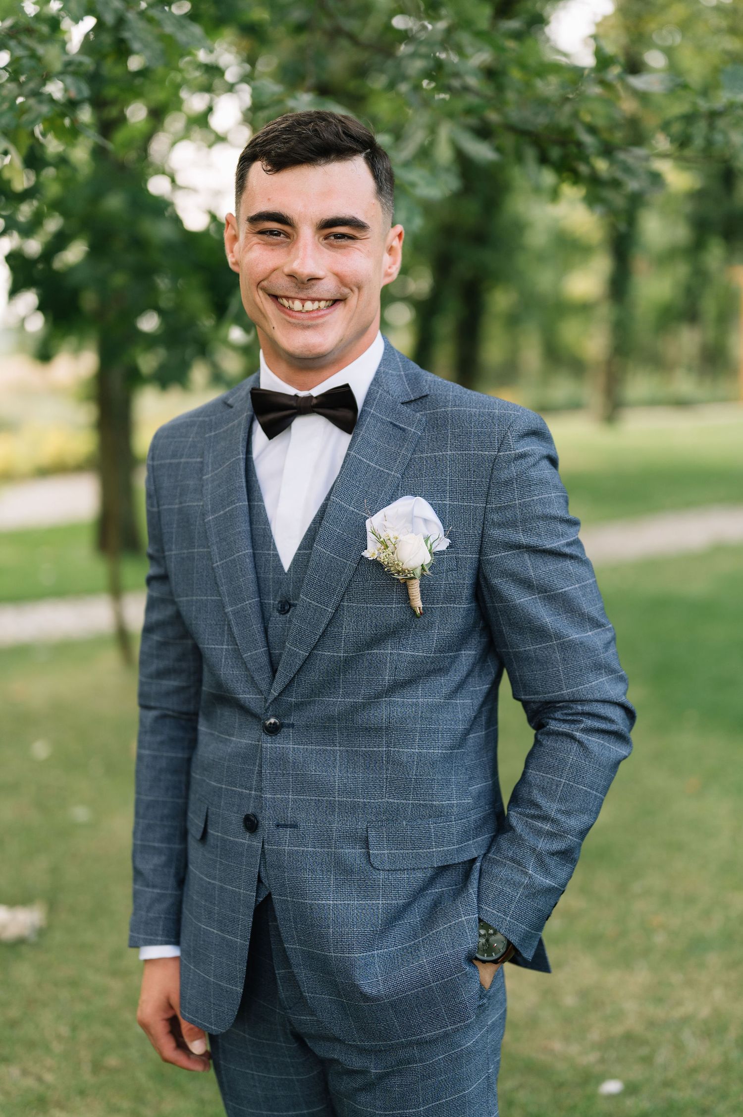 A groom in a grey windowpane suit with bowtie and white boutonniere poses outdoors in a garden setting.