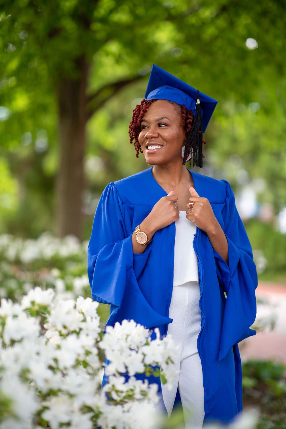Graduate in royal blue regalia posing near white flowers in garden setting.