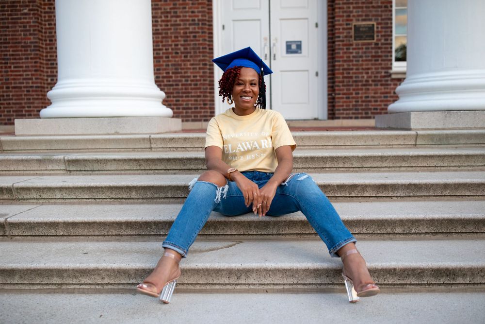 Graduate in casual attire sits casually on brick steps wearing blue graduation cap.