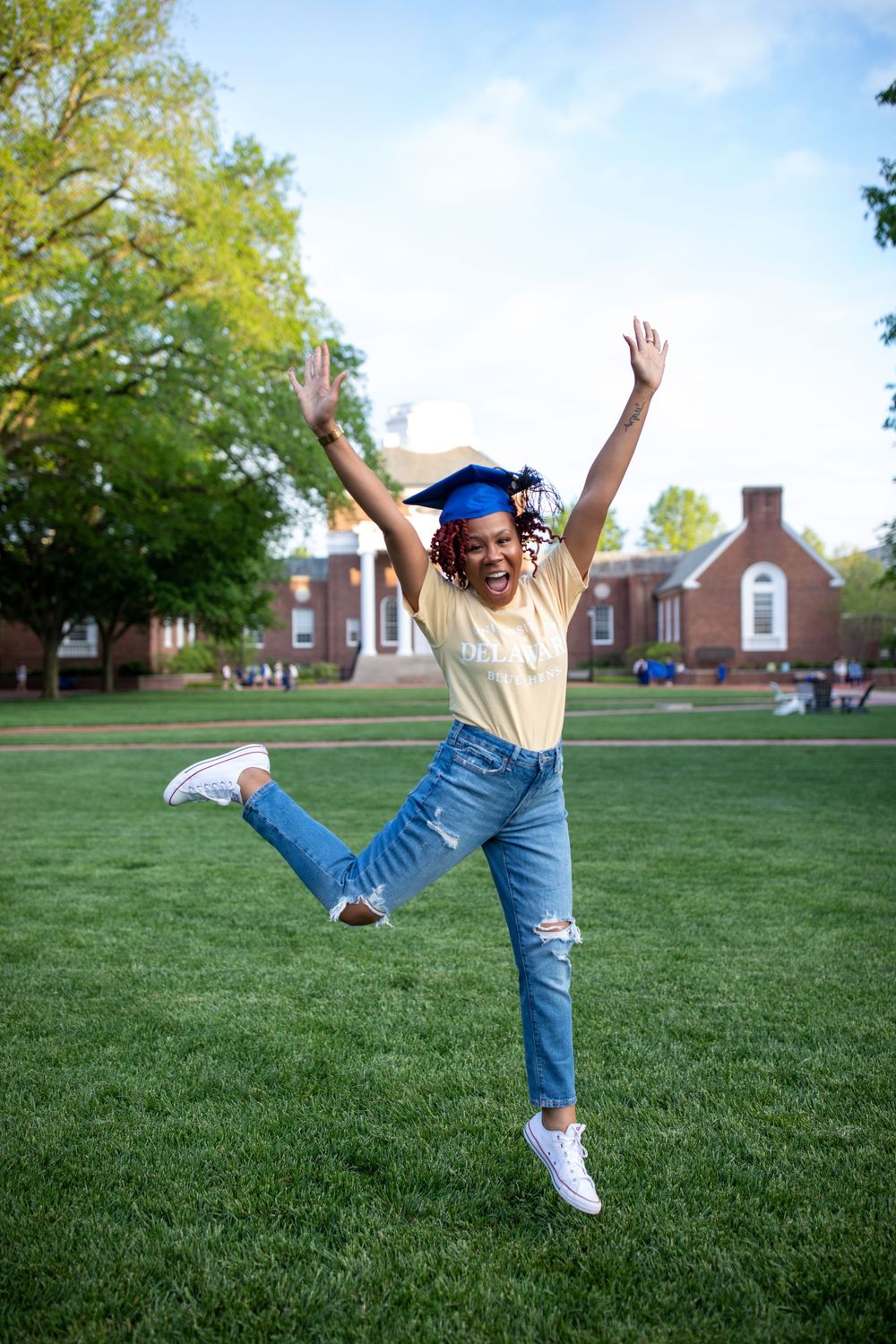 Graduate in casual attire and blue cap jumps joyfully on campus lawn.