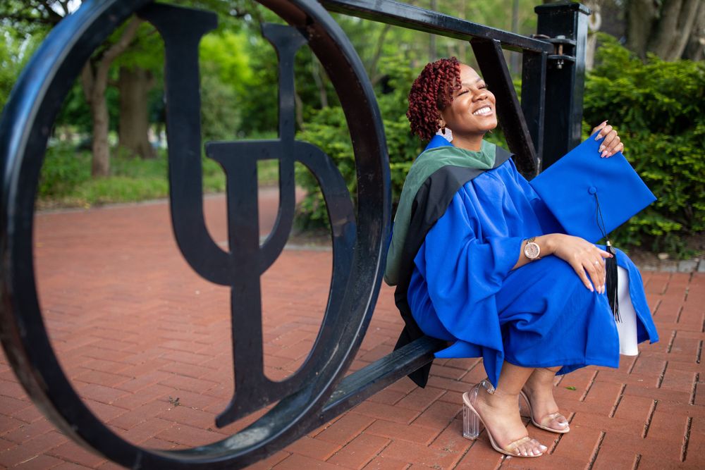 Graduate in white jumpsuit posing near university logo on campus.