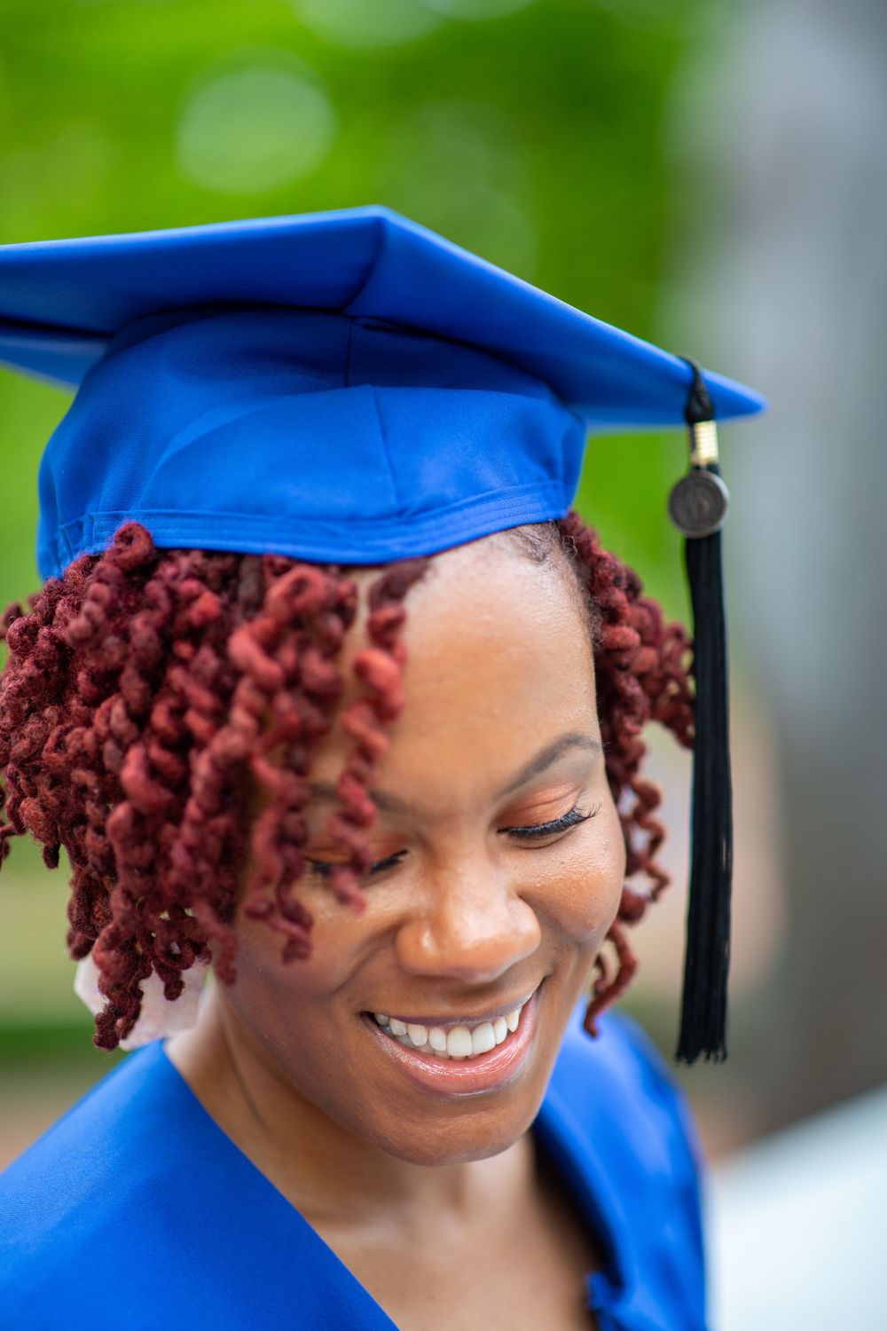 Graduate in blue cap and gown smiling against blurred green background.