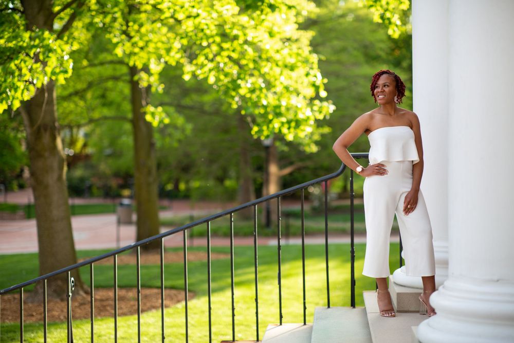 A graduate in an elegant white jumpsuit poses on a sunny porch with white columns and green trees in the background.