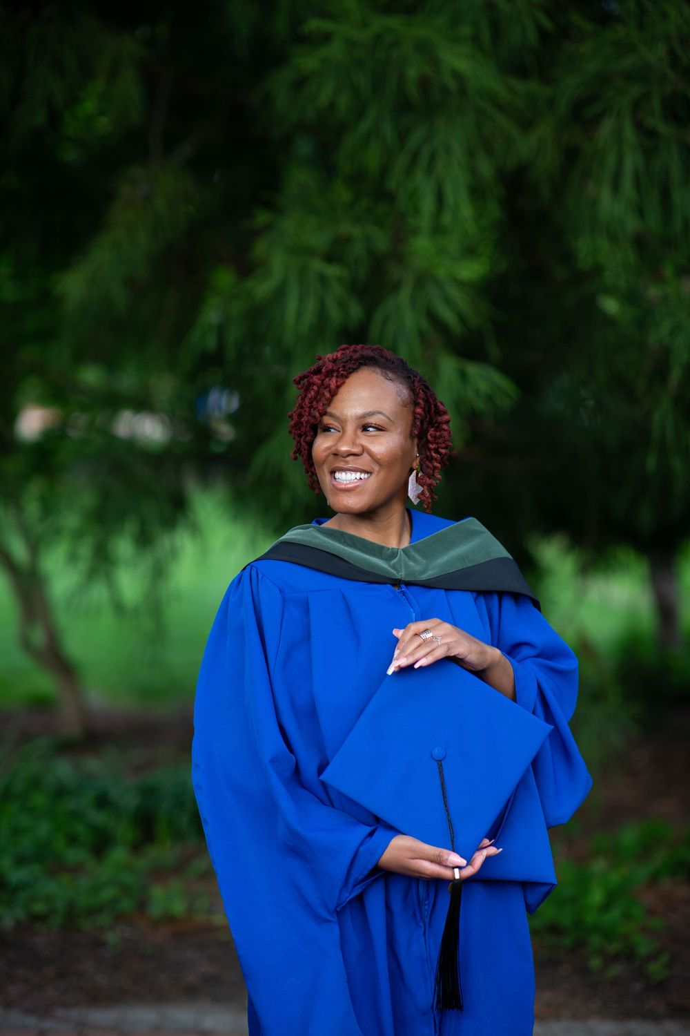 Graduate holding cap in blue academic regalia against green foliage background.