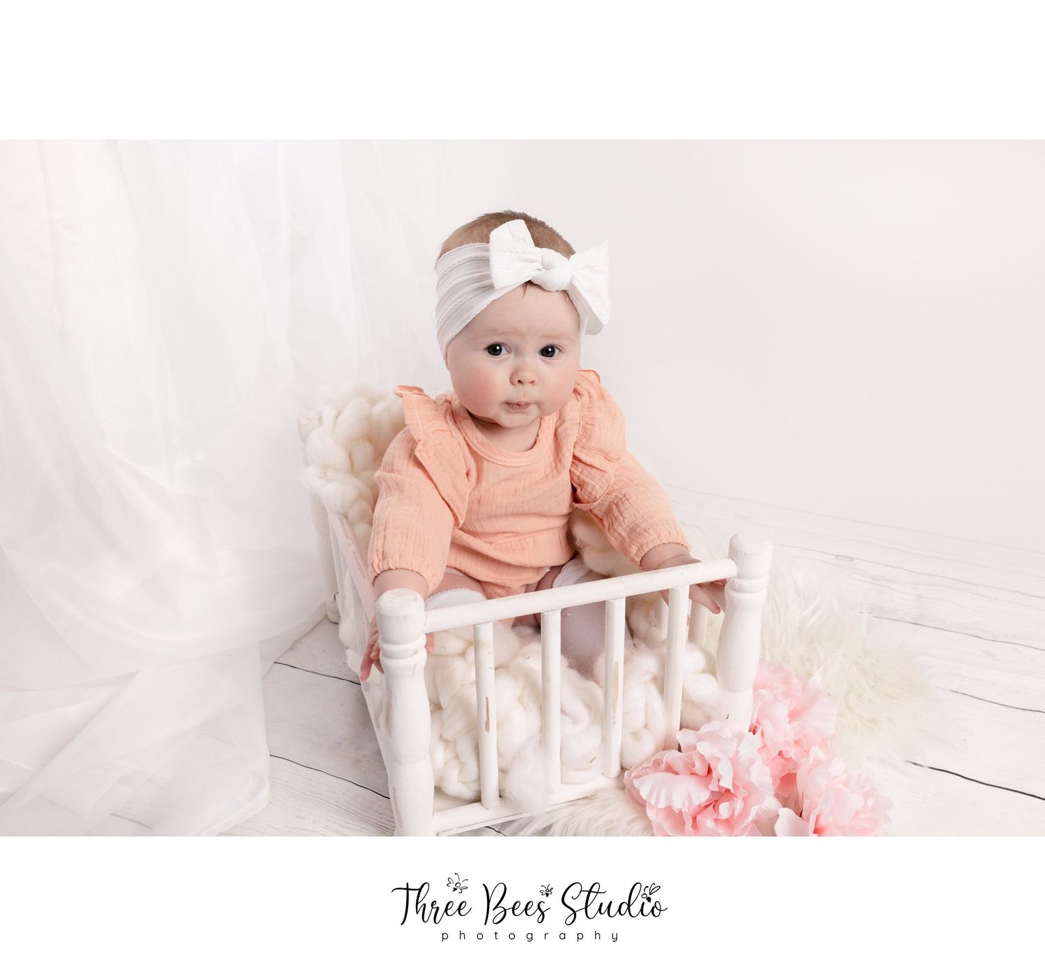 Baby in peachy outfit with white bow headband sitting in white wooden prop bed next to pink flowers.