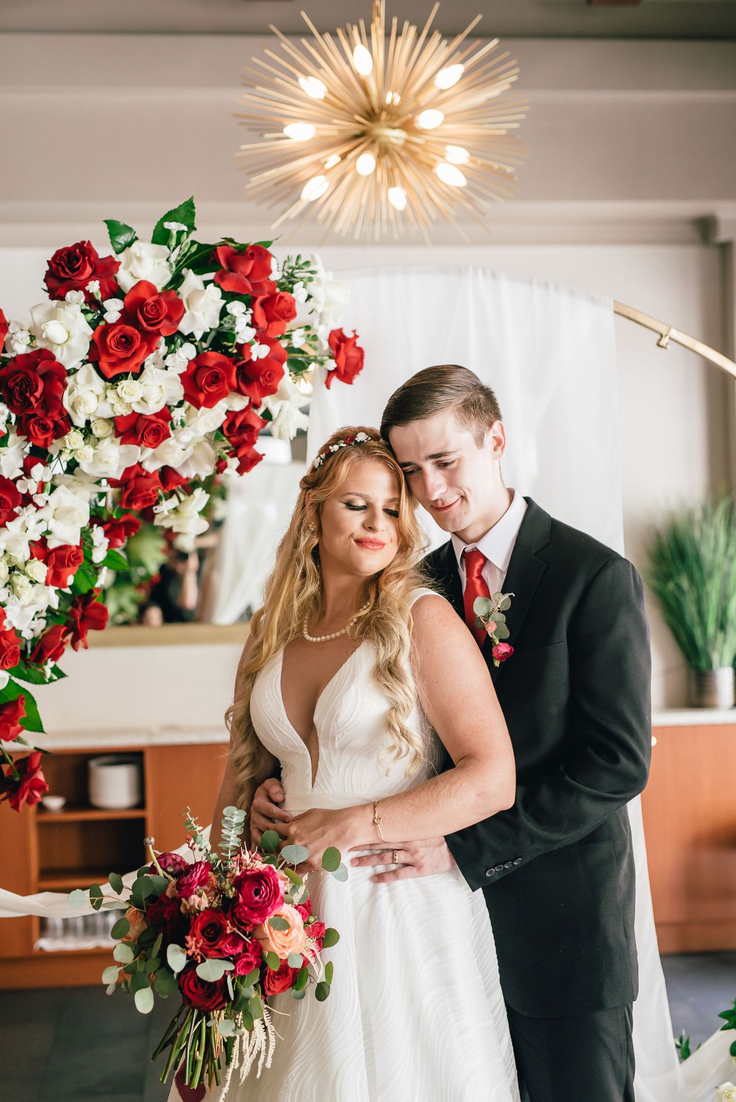 A romantic wedding moment under a starburst chandelier with red and white floral arrangements.