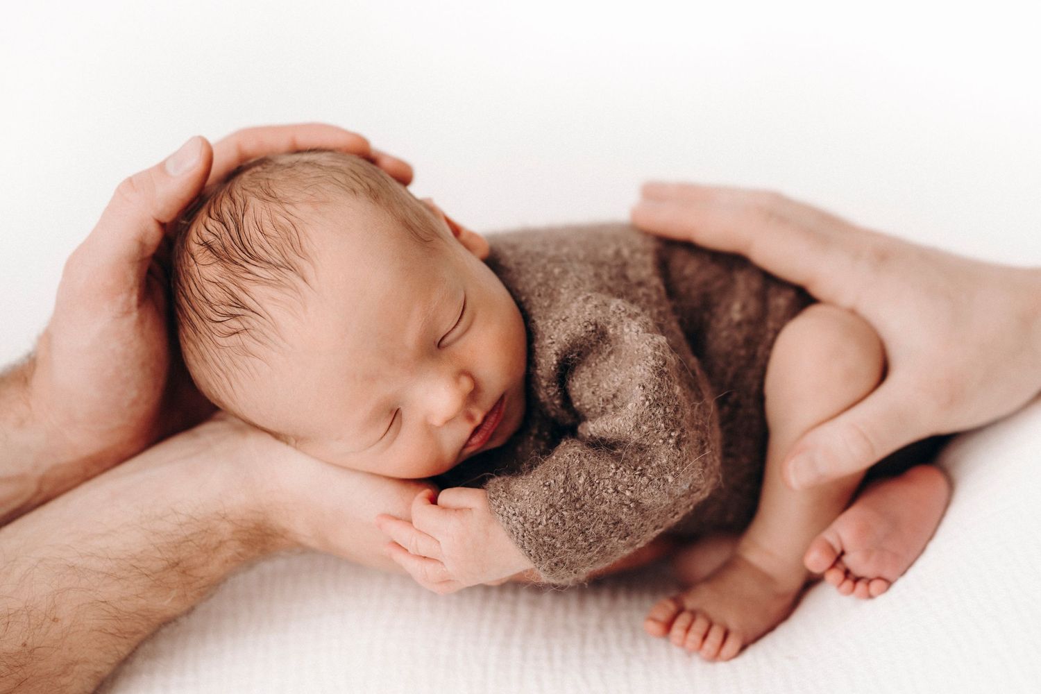 A peaceful newborn baby sleeping while wrapped in a brown blanket, supported by caring hands.