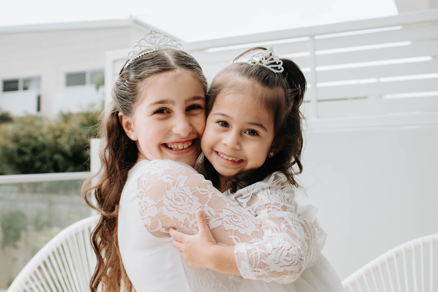 Two people in white lace dresses share a loving embrace outdoors.
