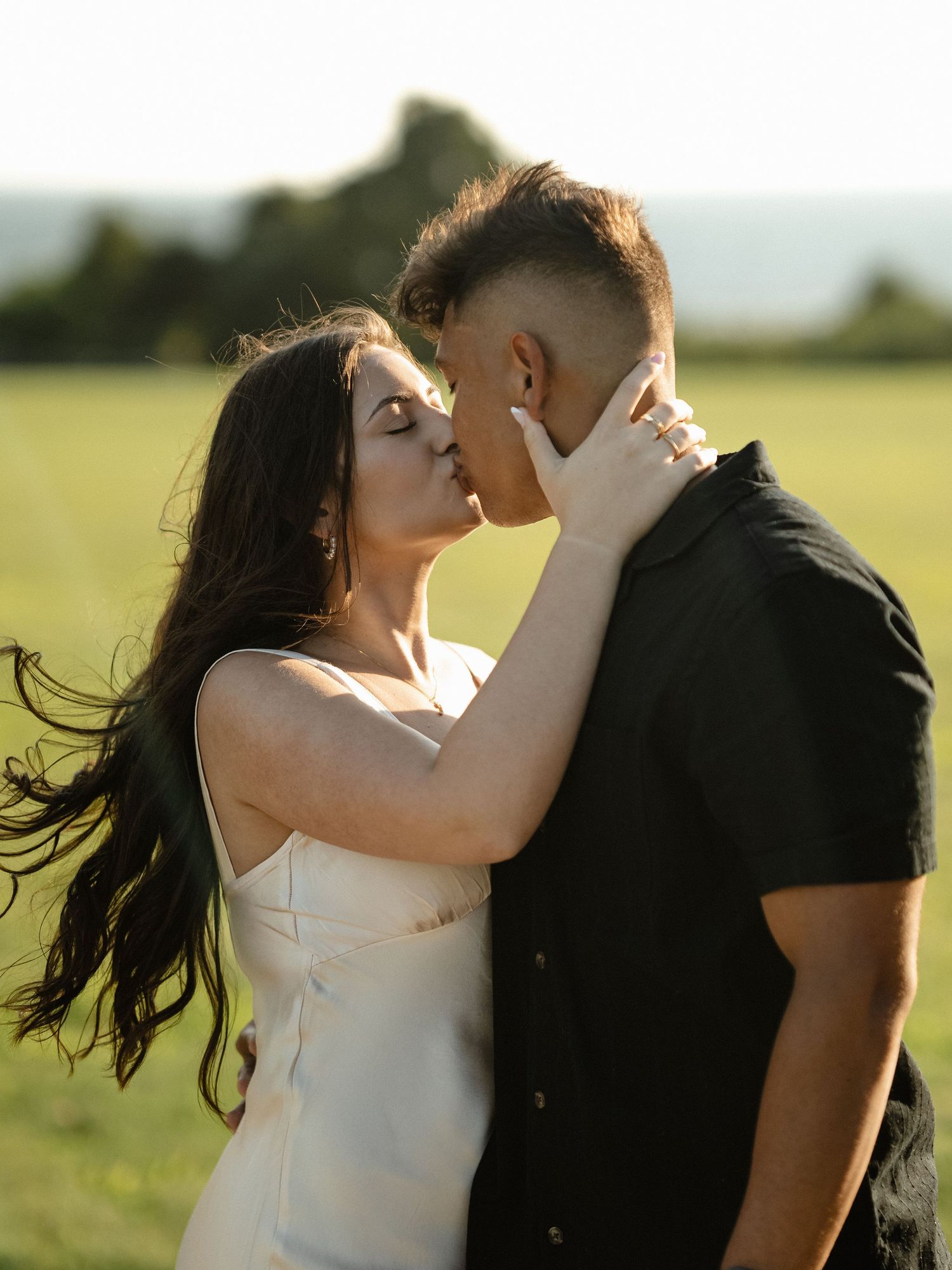 Couple sharing romantic kiss in sunlit field wearing contrasting black and white outfits.