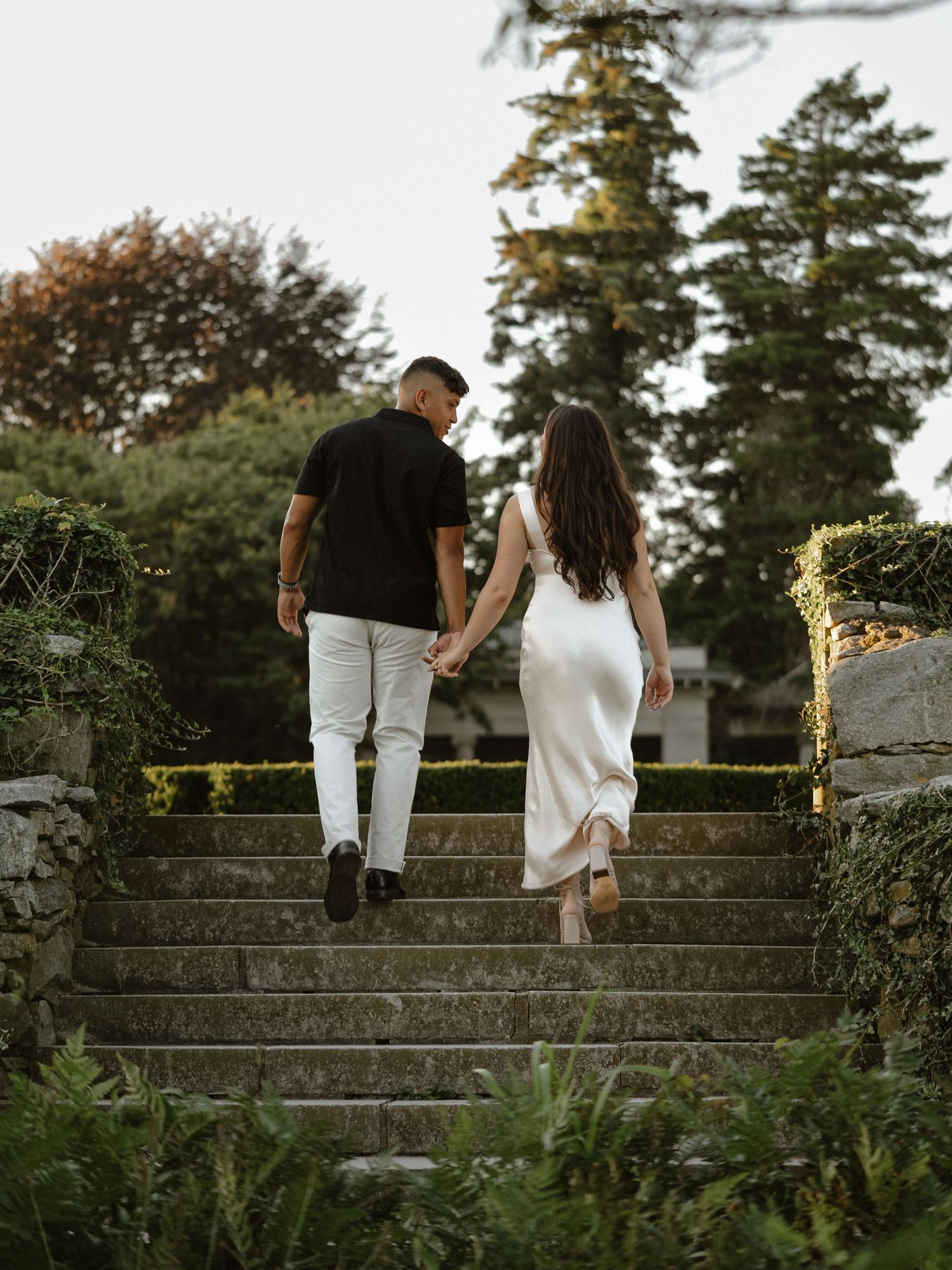 Couple walking down stone steps together in a garden at sunset.