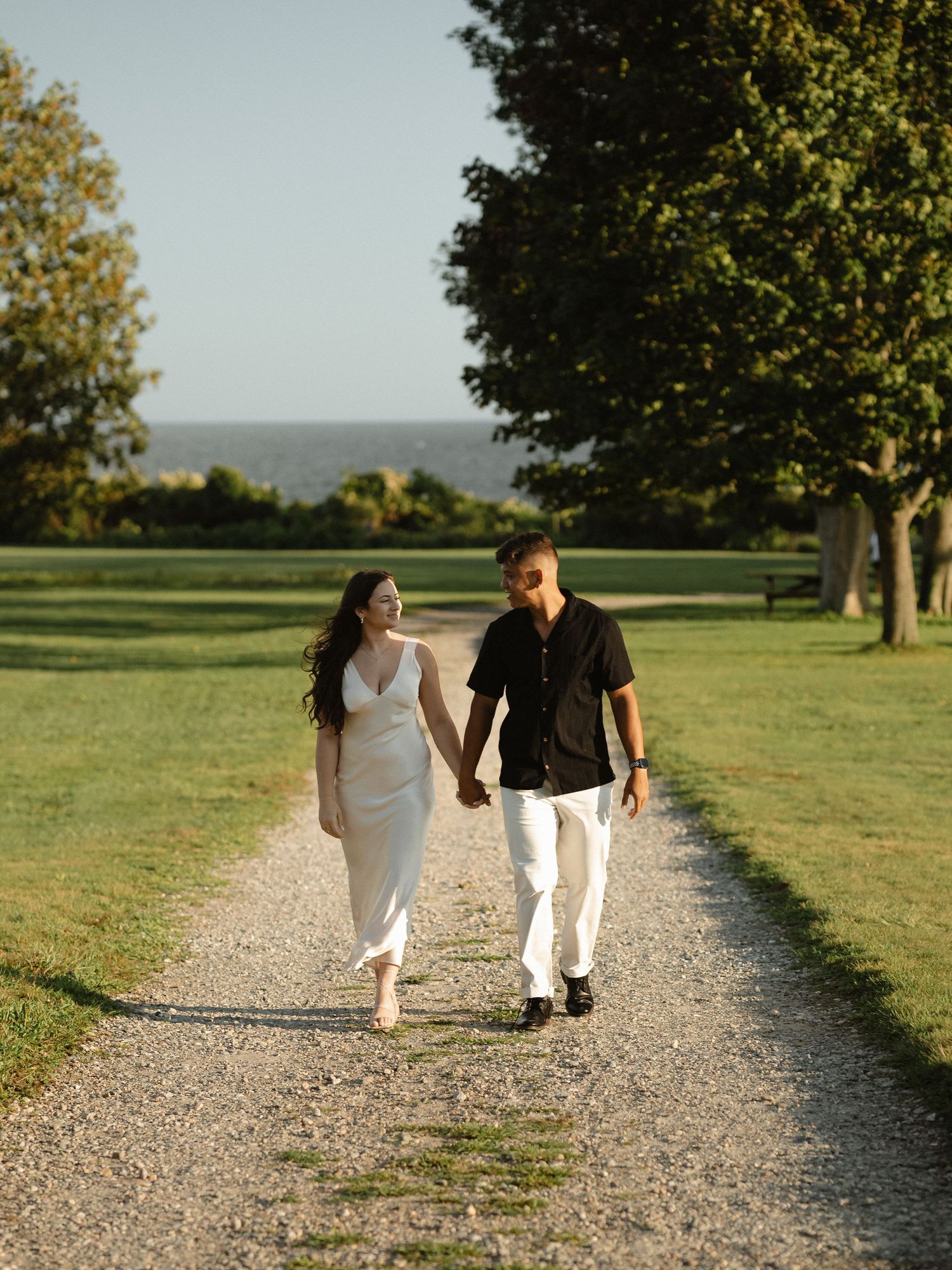 Couple walking hand in hand down a gravel path through green meadows.