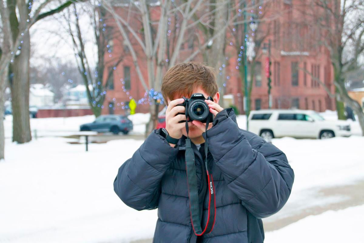Photographer wearing a blue winter coat takes pictures on a snowy winter day in front of a brick building.