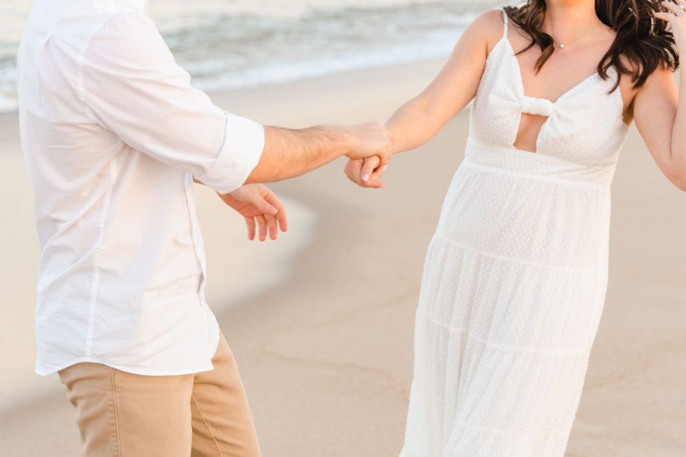 Couple holding hands on a sandy beach, woman in white maxi dress and man in white shirt and tan pants.