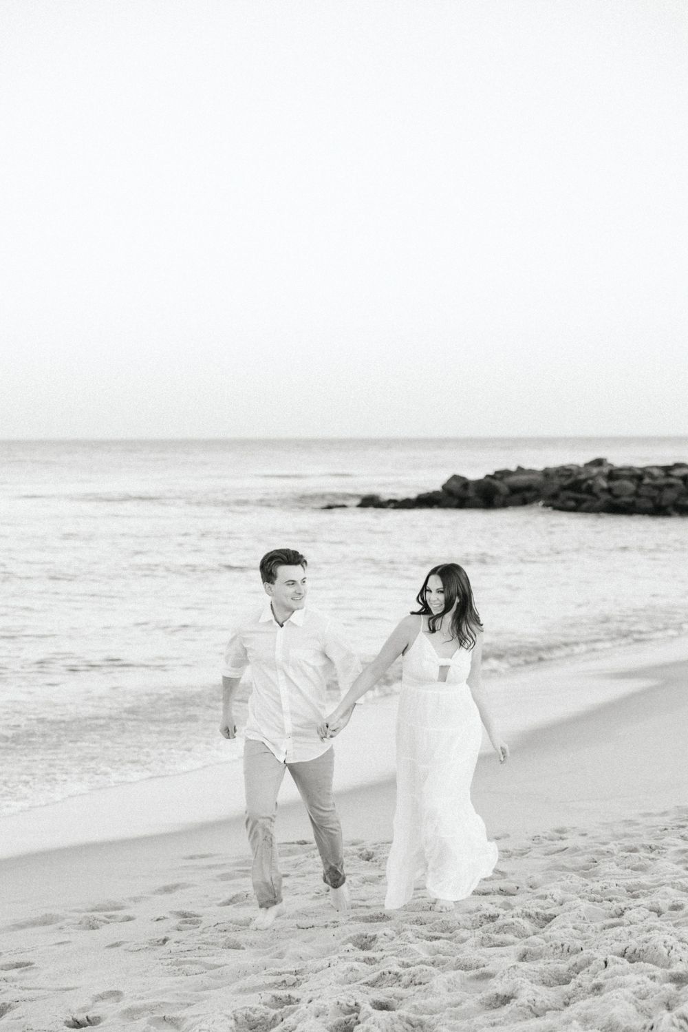Black and white photo of a couple walking hand in hand along the ocean shoreline at sunset.