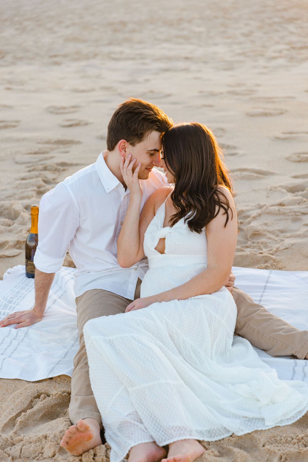 Couple sharing a romantic almost-kiss while seated on a beach blanket at sunset.