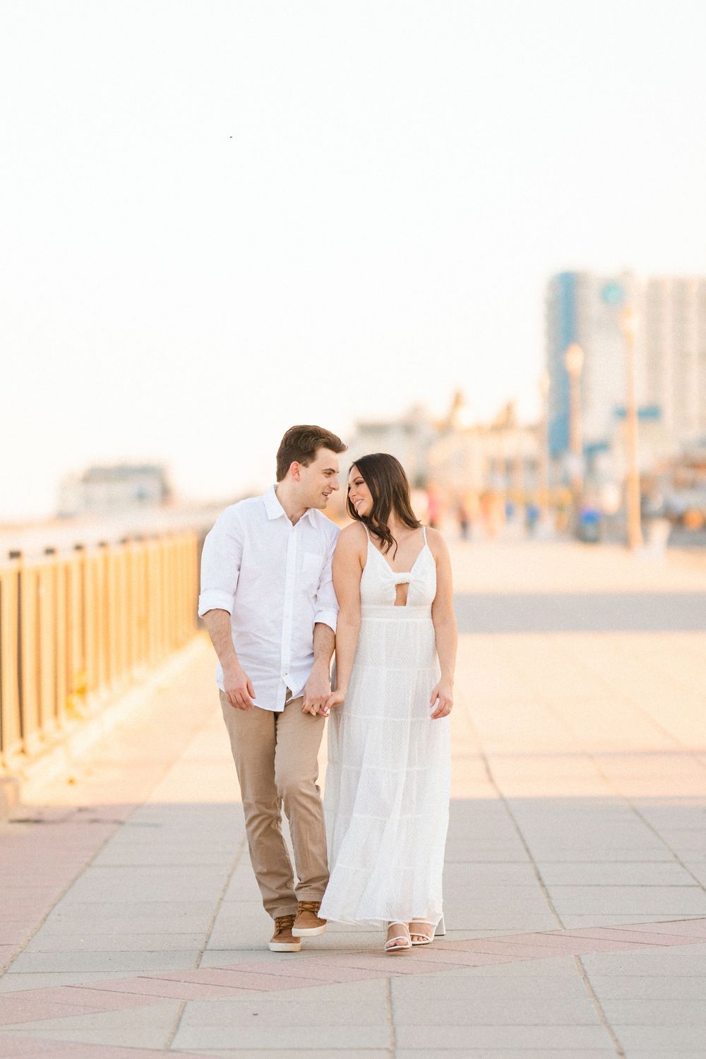 Couple walking and smiling at each other on a sunny boardwalk with city buildings in background.