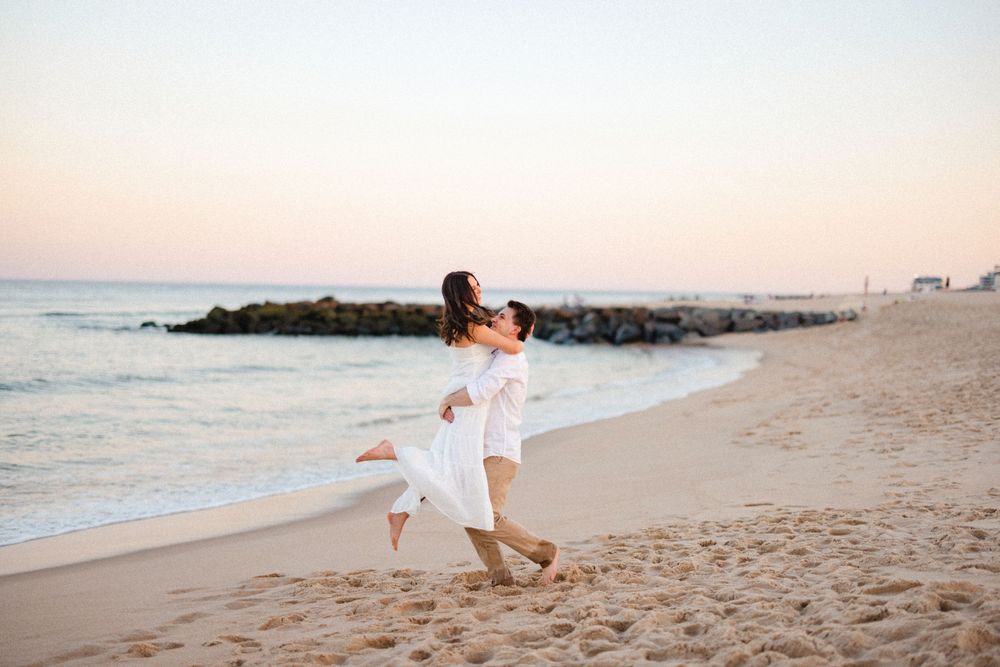 Man lifting woman in white dress on a sandy beach at sunset with rocky jetty in background.