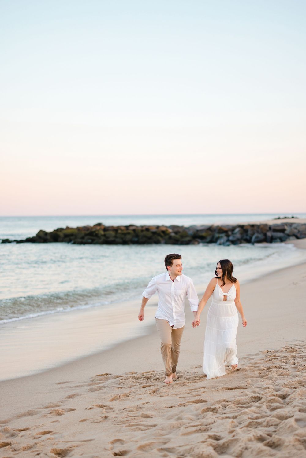 Couple walking hand in hand on a sandy beach at sunset, woman in white dress and man in white shirt.