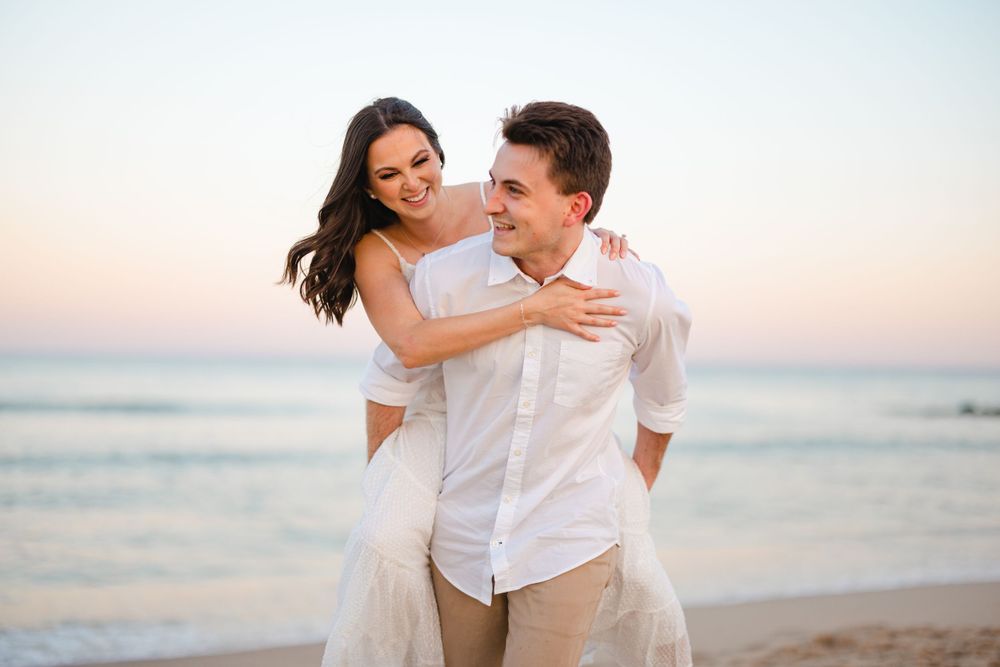 Happy couple embracing on a beach at sunset, woman in white dress hugging man from behind.