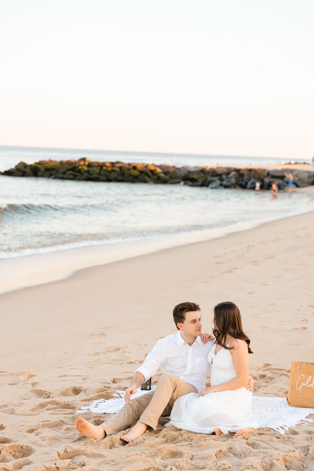 Couple sitting together on a beach blanket near the shoreline at sunset with rocky jetty behind them.