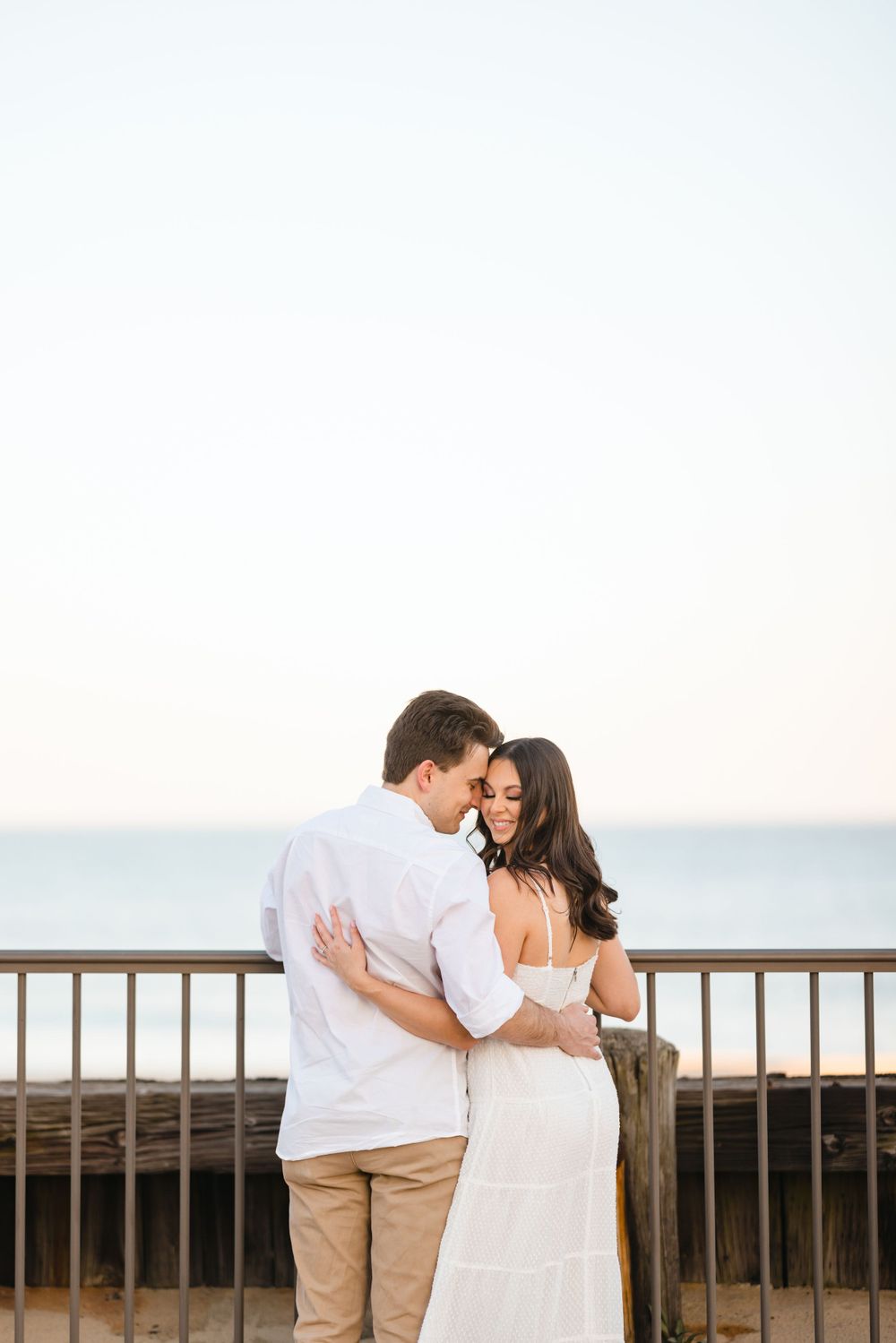 Couple embracing on a beachfront railing at sunset, man in white shirt and woman in white dress.