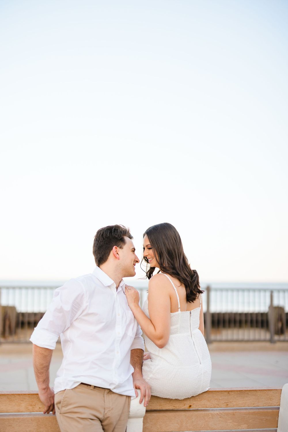 Couple leaning on a boardwalk railing smiling at each other with the ocean in the background.