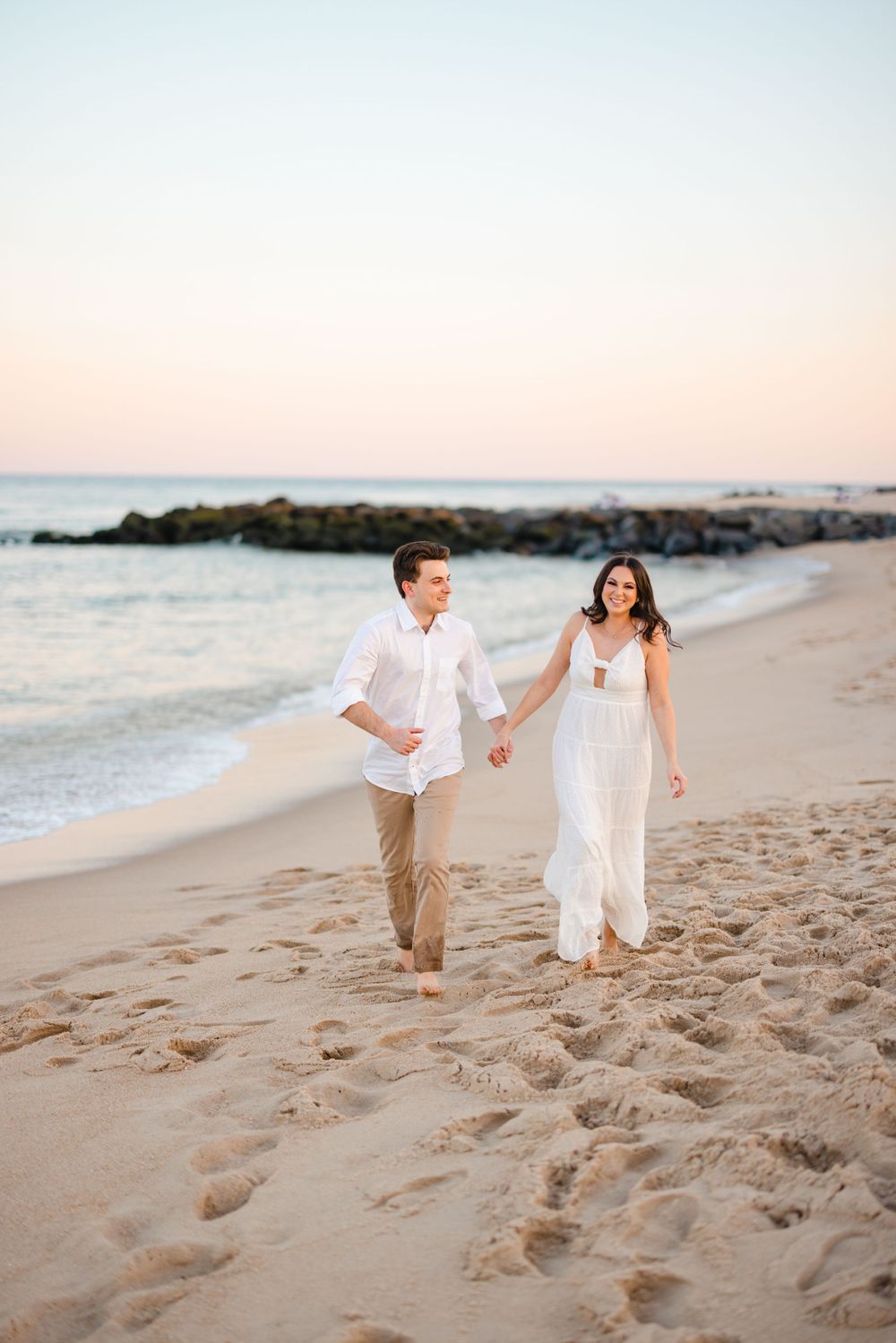 Smiling couple holding hands and strolling along a sandy beach at dusk near rocky jetty.