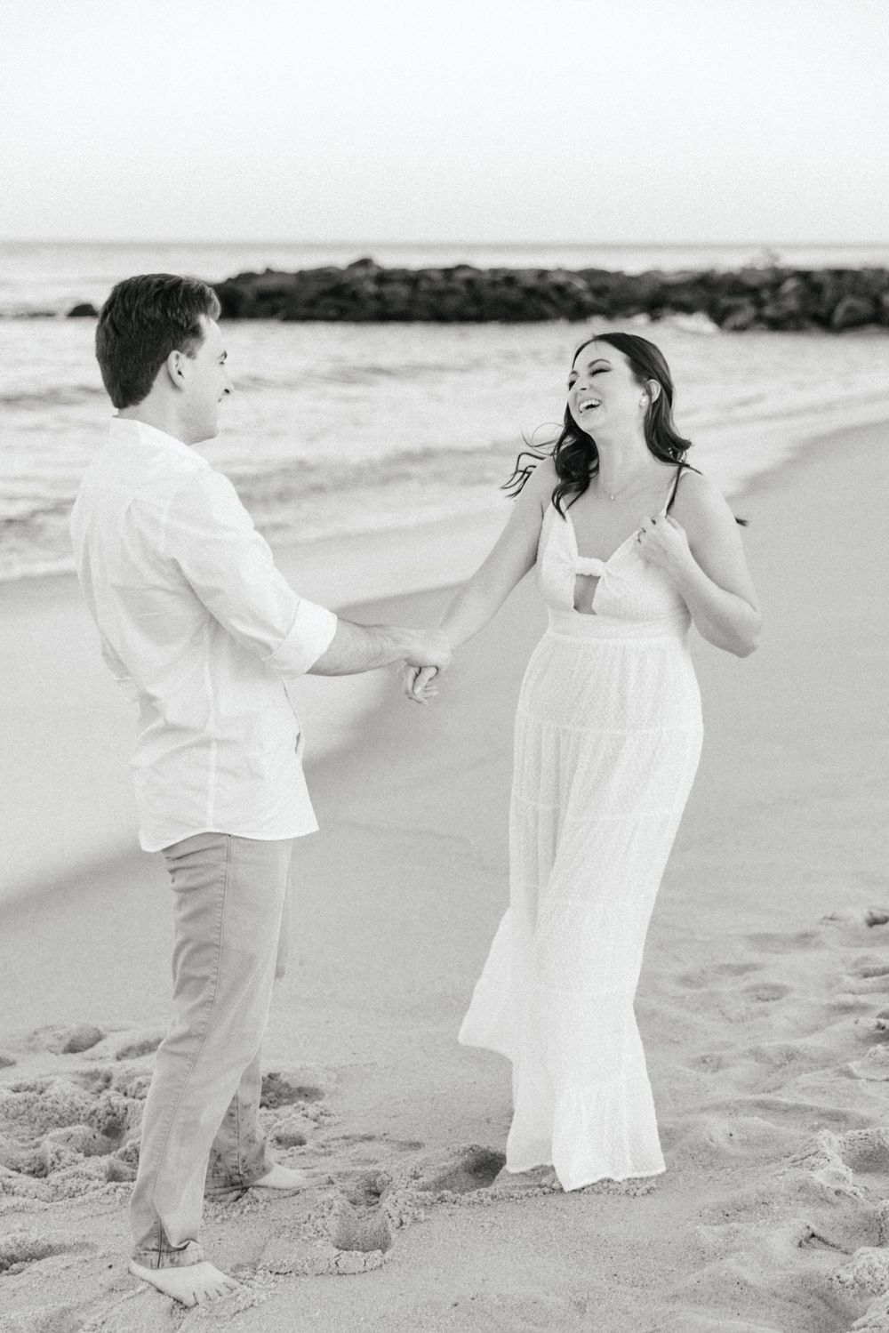 Black and white photo of a laughing couple holding hands and dancing on a sandy beach by the ocean.