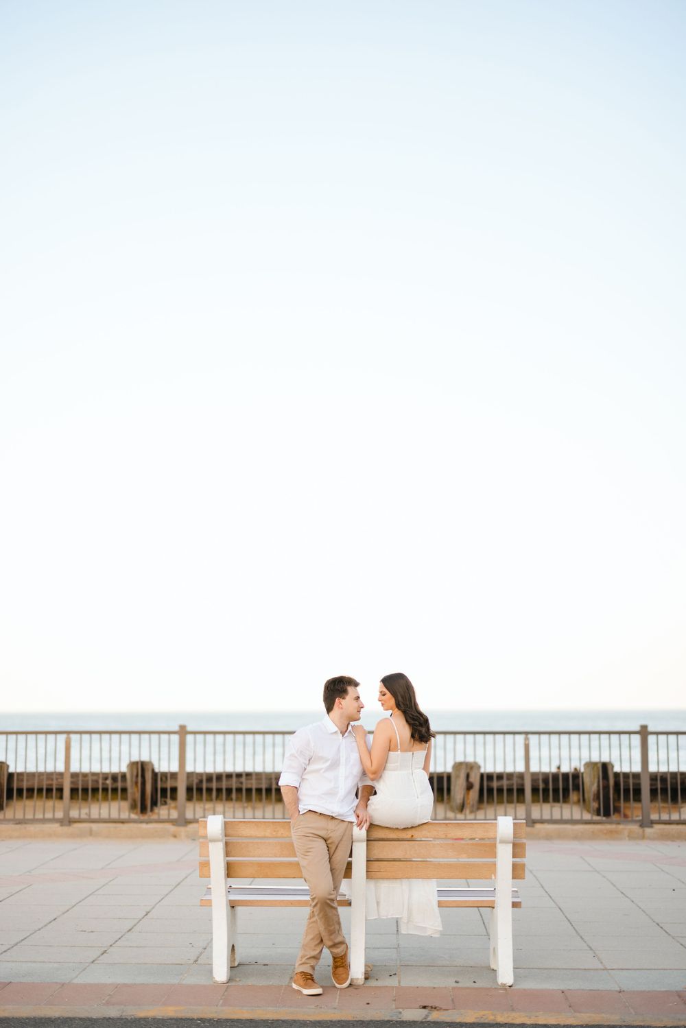 Couple sitting on a boardwalk bench facing each other with the ocean horizon in the background.