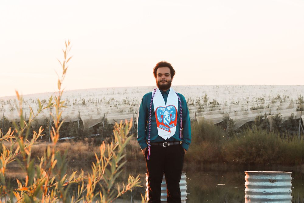 Man in turquoise graduation robe standing outdoors at sunset with sparse trees in background.